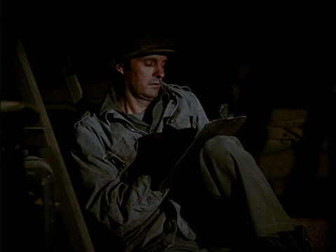 Hawkeye sitting under a table, wearing a helmet, writing on a pad of paper